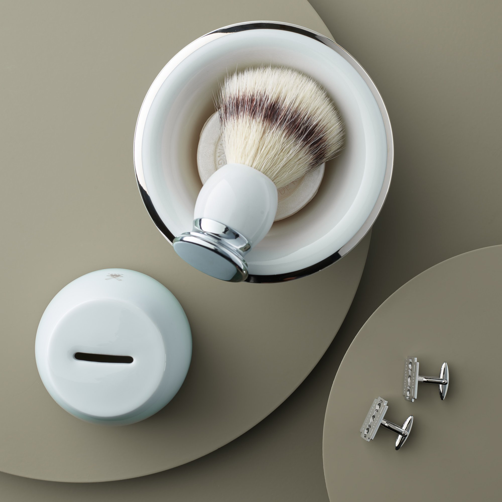 Ein Rasierpinsel in einer Schale mit Seife, eine weiße Rasierseifenschale mit Schlitz und zwei silberne Rasierklingenhalter auf einem grauen Hintergrund.